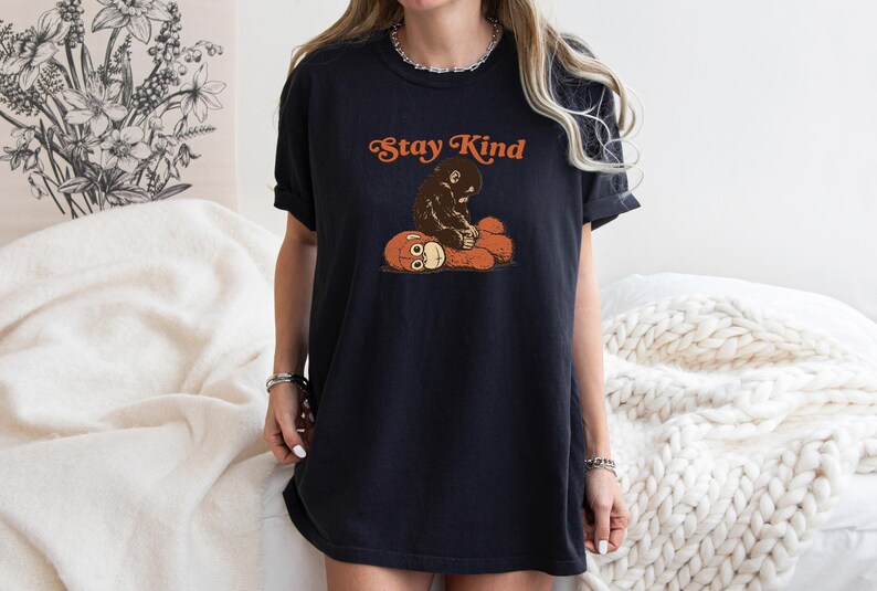 May include: Black t-shirt with the text "Stay Kind" in orange lettering. The shirt features a graphic of a brown ape hugging a teddy bear. The shirt is a relaxed fit.