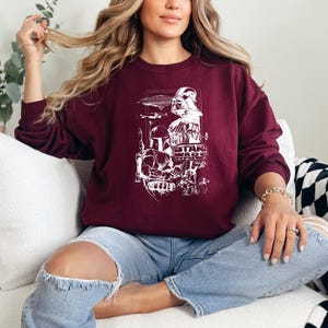 May include: Burgundy crewneck sweatshirt featuring a white graphic of Star Wars characters and the Star Wars logo. The design includes Darth Vader, Boba Fett, and other iconic elements from the film series. The sweatshirt is worn with blue jeans.