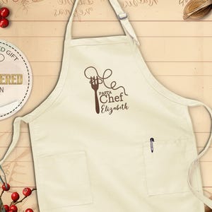 May include: Cream-colored apron with a brown graphic of a fork and pasta, the words "Pasta Chef Elizabeth" are below the graphic. The apron has two pockets and a pen in the right pocket. The apron is a personalized embroidered design.