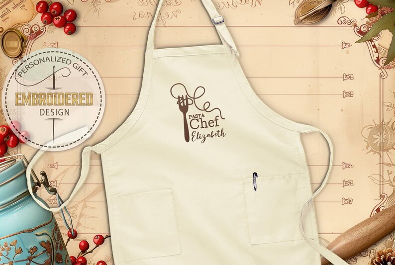 May include: Cream-colored apron with a brown graphic of a fork and pasta, the words "Pasta Chef Elizabeth" are below the graphic. The apron has two pockets and a pen in the right pocket. The apron is a personalized embroidered design.