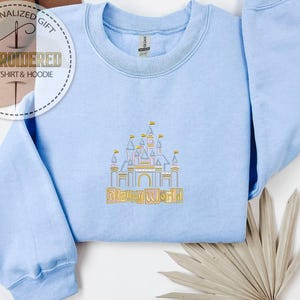 Puede incluir: Sudadera azul claro bordada con un dise&ntilde;o de castillo y la inscripci&oacute;n dorada "Disney World". La sudadera tiene cuello redondo y mangas largas. La imagen tambi&eacute;n incluye un logotipo de "Personalized Gift".