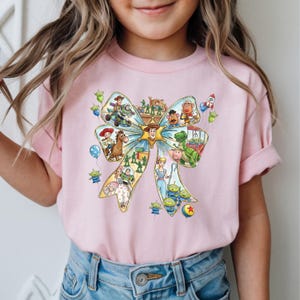 May include: A light pink t-shirt featuring a large bow design filled with various Toy Story characters. The bow is light blue and yellow, with characters like Woody, Buzz Lightyear, and the aliens. The shirt is worn by a person.