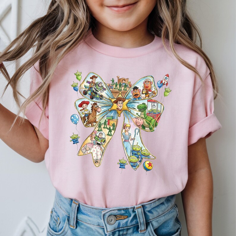 May include: A light pink t-shirt featuring a large bow design filled with various Toy Story characters. The bow is light blue and yellow, with characters like Woody, Buzz Lightyear, and the aliens. The shirt is worn by a person.