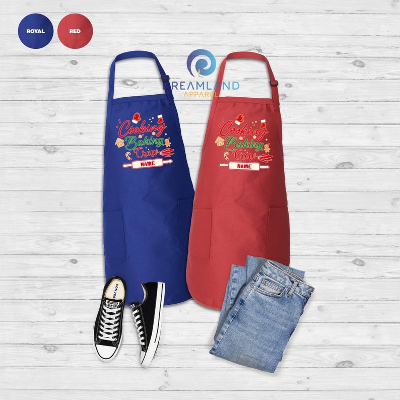 Może przedstawiać: Dwa świąteczne fartuchy, jeden w kolorze kr&oacute;lewskim, a drugi czerwony, z napisem "Cooking Baking Crew" i miejscem na imię. Fartuchy mają kieszeń z przodu.
