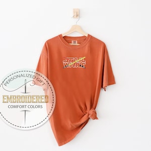 Może przedstawiać: Koszulka w kolorze rdzy z kr&oacute;tkimi rękawami i haftowanym wzorem w kolorze białym z napisem "STAR WARS" i dwoma skrzyżowanymi mieczami świetlnymi za tekstem.