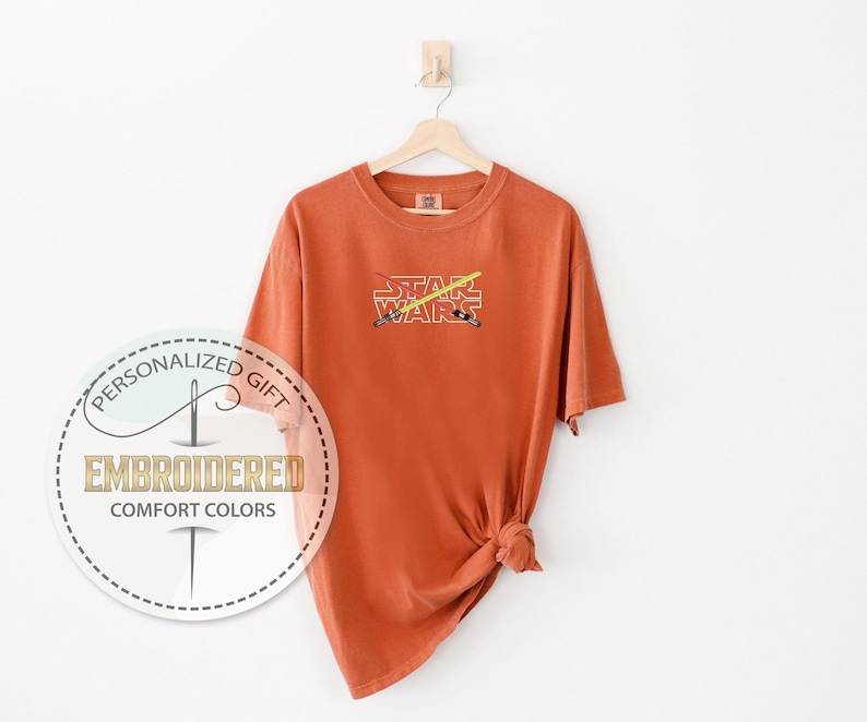 Może przedstawiać: Koszulka w kolorze rdzy z kr&oacute;tkimi rękawami i haftowanym wzorem w kolorze białym z napisem "STAR WARS" i dwoma skrzyżowanymi mieczami świetlnymi za tekstem.