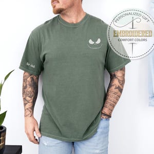 K&ouml;nnte beinhalten: Olivgr&uuml;nes T-Shirt mit einem wei&szlig;en, gestickten Jack Skellington-Gesicht auf der Brust. Der &Auml;rmel hat den gestickten Text "Her Jack". Das Shirt hat eine lockere Passform und ist mit "Embroidered Comfort Colors" gekennzeichnet.