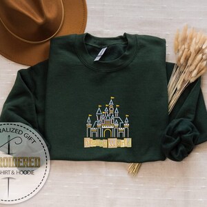 Puede incluir: Sudadera verde oscuro con un dise&ntilde;o de castillo bordado y las palabras "Disney World" en dorado. La sudadera est&aacute; doblada, con un sombrero marr&oacute;n y flores secas en el fondo. Tambi&eacute;n se ven las palabras "Embroidered Sweatshirt & Hoodie".