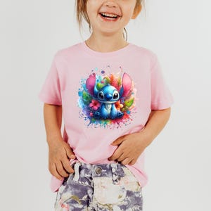 Op de afbeelding: Lichtroze T-shirt met een kleurrijke afbeelding van een blauw stripfiguur met grote oren, omringd door bloemen en verfspatten. Het t-shirt heeft korte mouwen en is geschikt voor kinderen.