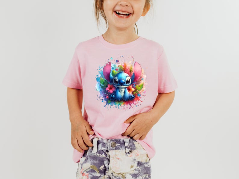 Op de afbeelding: Lichtroze T-shirt met een kleurrijke afbeelding van een blauw stripfiguur met grote oren, omringd door bloemen en verfspatten. Het t-shirt heeft korte mouwen en is geschikt voor kinderen.