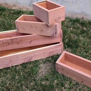 Puede incluir: Una colección de jardineras rectangulares de madera sin terminar de varios tamaños. Las jardineras están hechas de madera de color marrón claro y están apiladas sobre una superficie cubierta de hierba. Las jardineras están vacías y listas para usar.