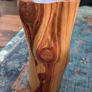 May include: A cylindrical wooden stool crafted from a natural log, featuring a smooth, polished top surface and a vertical grain pattern. The exterior displays prominent knots and a warm, variegated color palette ranging from light tan to deep reddish-brown.