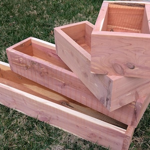 Peut inclure: Quatre jardinières rectangulaires en bois non fini de tailles différentes sont empilées sur de l'herbe verte. Les jardinières sont en bois de couleur claire avec des veines et des nœuds visibles. L'arrangement suggère un affichage en gradins.