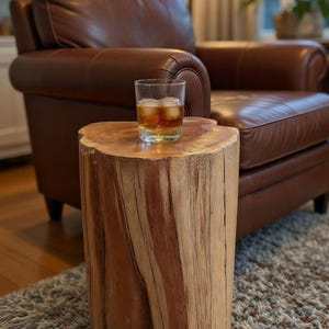 May include: A natural wood side table with a glass of whiskey and ice. The table is cylindrical, with a rough-hewn, textured surface. The table is approximately 18 inches tall and sits on a patterned rug. A brown leather armchair is in the background.