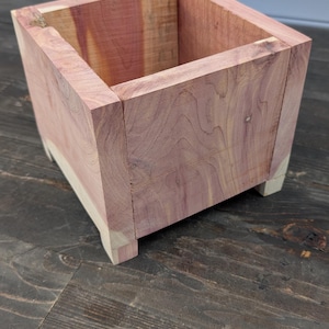 Puede incluir: Una jardinera cuadrada de madera de color marrón rojizo, con vetas visibles y patas más claras. La jardinera está diseñada para uso en interiores o exteriores y mide unos 20 cm de alto.