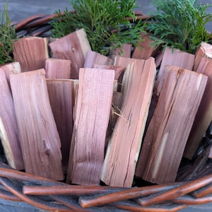 Puede incluir: Una cesta de mimbre llena de piezas rectangulares de madera de cedro y ramitas de follaje verde. Las piezas de cedro tienen un tono marrón rojizo con vetas de madera visibles. La cesta es de material tejido marrón oscuro.