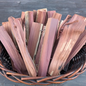 Puede incluir: Una cesta de mimbre marrón llena de trozos de madera de cedro de color marrón rojizo. La madera tiene un patrón de vetas natural y está dispuesta en forma de abanico. La cesta tiene un asa.