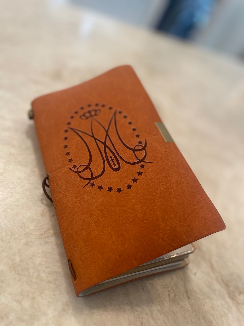 May include: A brown leather-bound journal with a decorative embossed design featuring a monogram and a cross. The journal has a silver clasp and a frayed brown string.