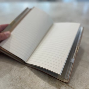 May include: A brown leather-bound journal with lined pages is open and lying on a light gray surface. The journal has a clear plastic pocket on the back cover.
