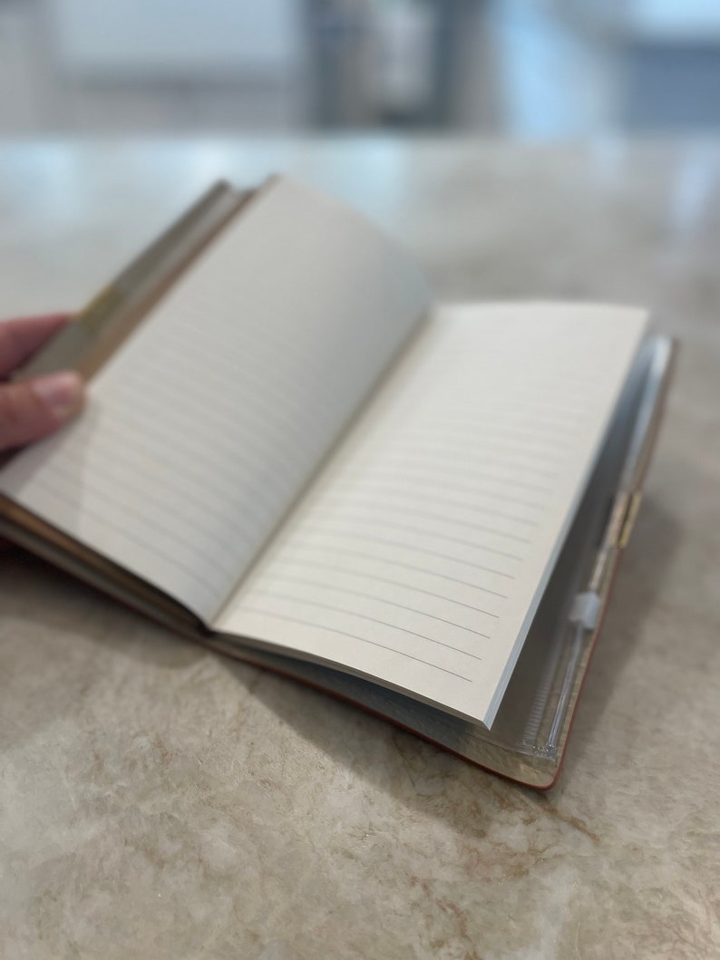 May include: A brown leather-bound journal with lined pages is open and lying on a light gray surface. The journal has a clear plastic pocket on the back cover.