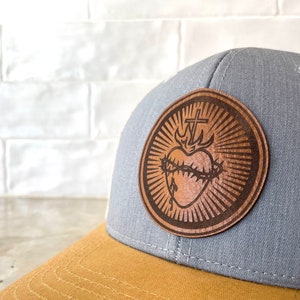 May include: A gray and brown trucker hat with a leather patch featuring a stylized heart with a crown of thorns and a cross. The patch is brown with a light brown background.