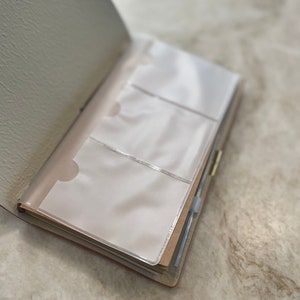 May include: Open beige leather journal with three clear plastic pockets for holding cards or photos.