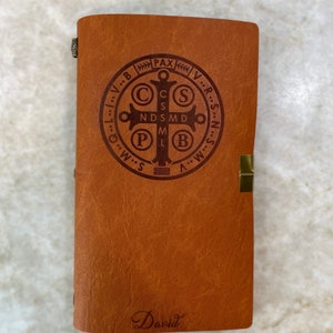 May include: A brown leather journal with a gold clasp and a metal inscription of the Saint Benedict Medal. The inscription reads "V.R.S.N.S.M.D.P.B.V.R.S.N.S.M.D.P.B. PAX" and the journal is signed "David".