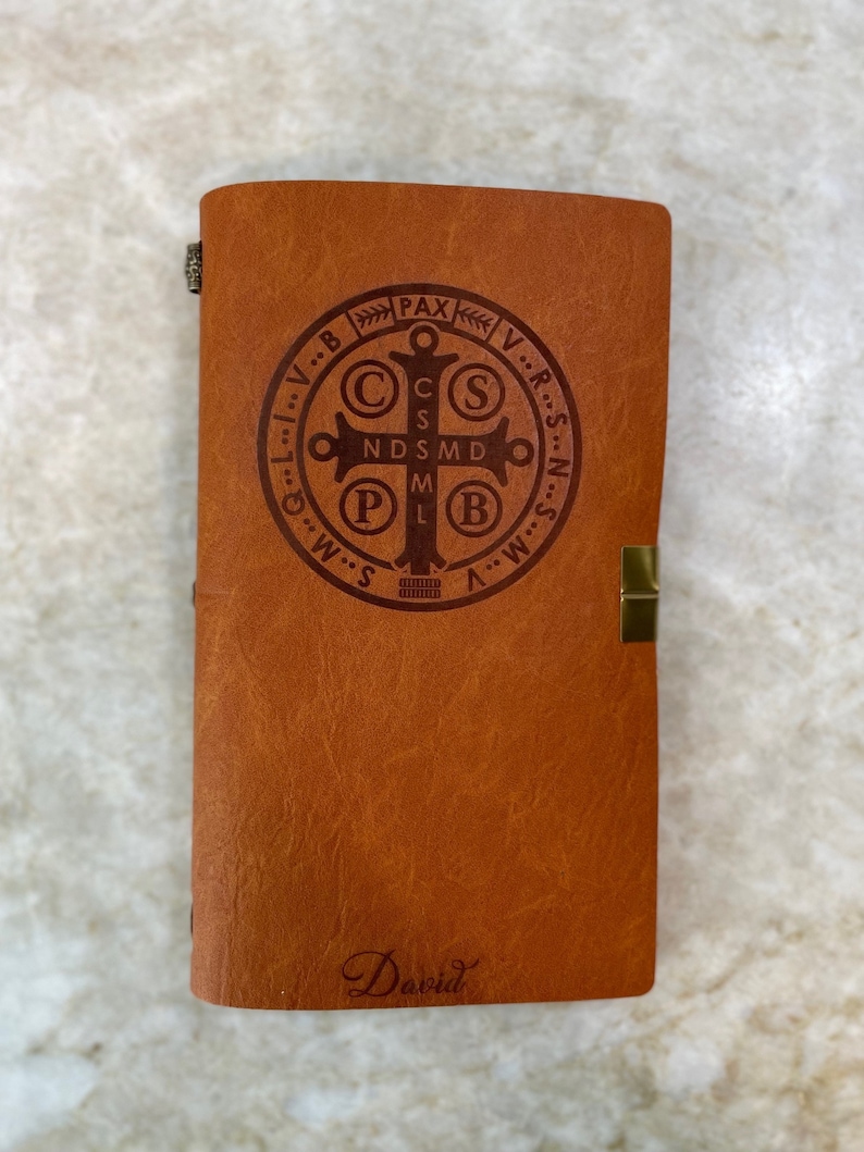 May include: A brown leather journal with a gold clasp and a metal inscription of the Saint Benedict Medal. The inscription reads "V.R.S.N.S.M.D.P.B.V.R.S.N.S.M.D.P.B. PAX" and the journal is signed "David".