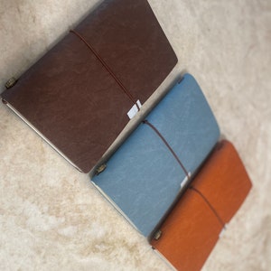May include: Three leather-bound journals in brown, blue, and orange. Each journal has a string closure and a textured cover.