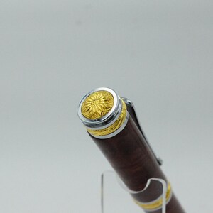 May include: Close-up of a brown pen with gold and silver accents. The pen's cap features a gold floral design surrounded by a silver band. The pen's body is a dark brown color with gold bands near the tip.