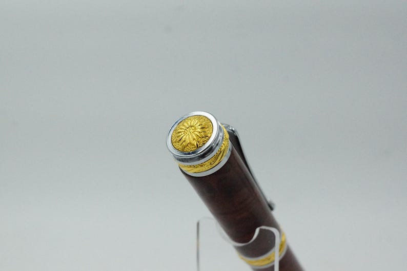 May include: Close-up of a brown pen with gold and silver accents. The pen's cap features a gold floral design surrounded by a silver band. The pen's body is a dark brown color with gold bands near the tip.