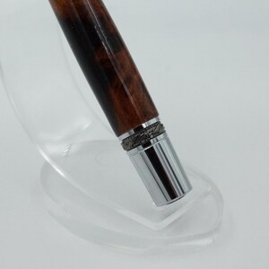May include: A close-up of a pen with a dark brown wooden barrel and a silver metal tip. The pen has a decorative silver band near the tip. The pen is displayed on a clear, curved stand.