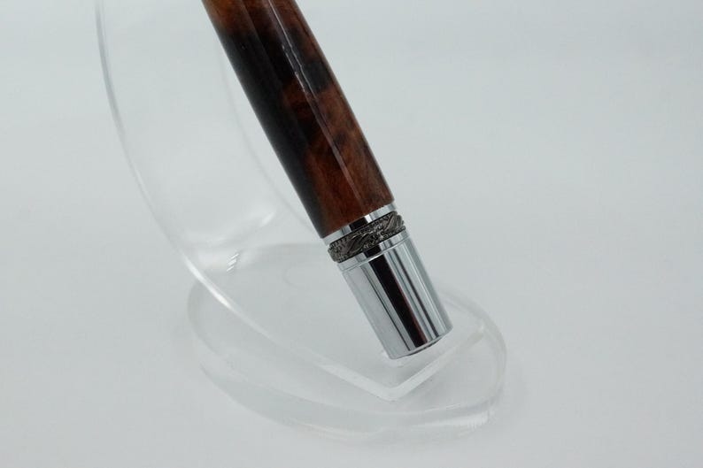 May include: A close-up of a pen with a dark brown wooden barrel and a silver metal tip. The pen has a decorative silver band near the tip. The pen is displayed on a clear, curved stand.