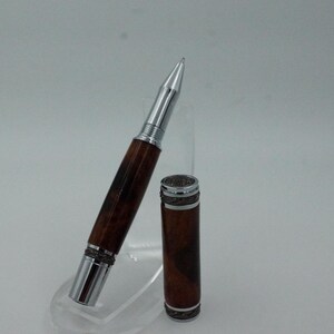 May include: A handcrafted pen with a wooden barrel and silver-colored metal accents. The pen is shown with its cap removed, revealing the writing tip. The wood grain pattern is visible on the barrel, and the pen is displayed on a clear stand.