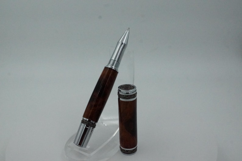 May include: A handcrafted pen with a wooden barrel and silver-colored metal accents. The pen is shown with its cap removed, revealing the writing tip. The wood grain pattern is visible on the barrel, and the pen is displayed on a clear stand.