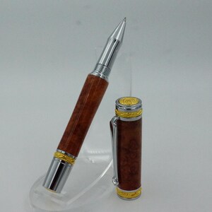 May include: A luxury pen with a silver tip and accents, a brown wooden barrel, and gold-colored trim. The pen's cap is detached and placed next to the pen. The pen is displayed on a clear stand.
