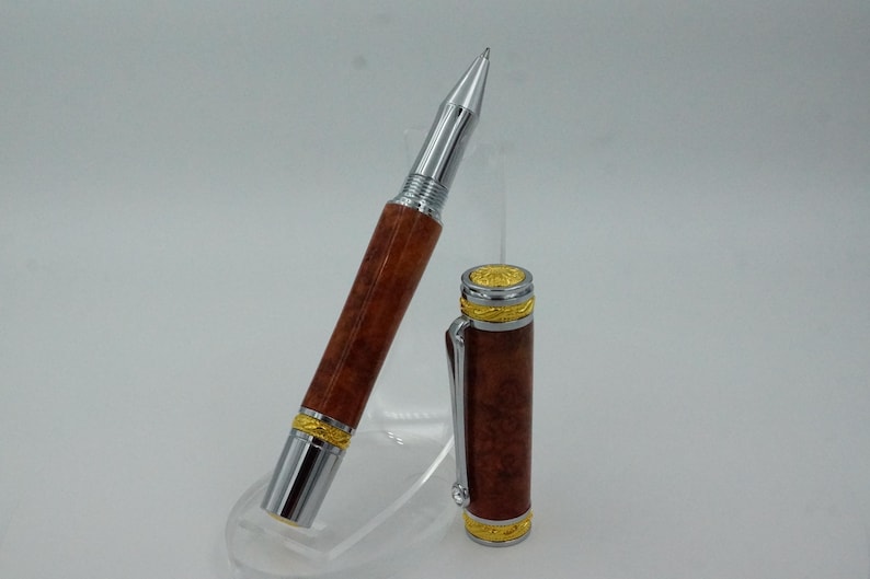 May include: A luxury pen with a silver tip and accents, a brown wooden barrel, and gold-colored trim. The pen's cap is detached and placed next to the pen. The pen is displayed on a clear stand.