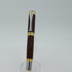 May include: A brown and silver pen with gold accents. The pen has a dark brown barrel, silver clip, and gold bands at the top and bottom. A small, clear gem is set near the top. The pen is displayed on a clear stand.