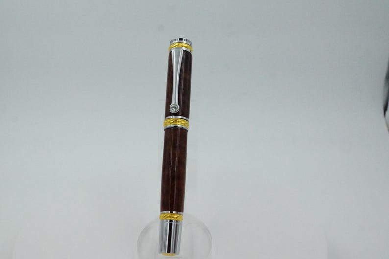 May include: A brown and silver pen with gold accents. The pen has a dark brown barrel, silver clip, and gold bands at the top and bottom. A small, clear gem is set near the top. The pen is displayed on a clear stand.