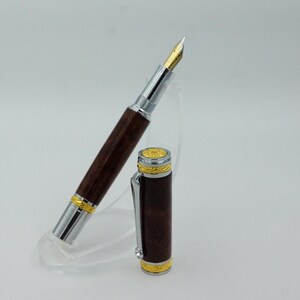 May include: A luxury fountain pen with a dark brown wooden barrel and silver accents. The pen features a gold nib and gold-colored trim. The cap is detached and placed next to the pen. The pen is displayed on a clear stand.