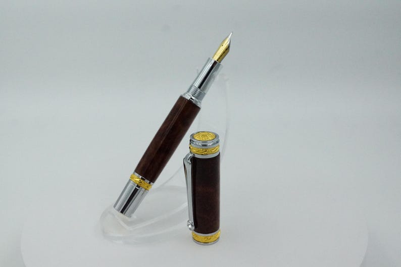 May include: A luxury fountain pen with a dark brown wooden barrel and silver accents. The pen features a gold nib and gold-colored trim. The cap is detached and placed next to the pen. The pen is displayed on a clear stand.