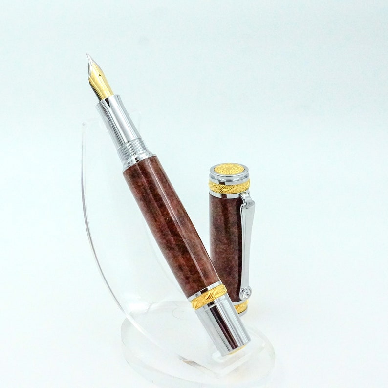 May include: A luxury fountain pen with a brown marbled barrel, silver accents, and a gold nib. The cap is off, revealing the pen's design. The pen is displayed on a clear stand, showcasing its elegant details.