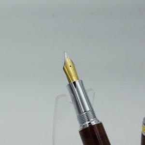 May include: Close-up of a fountain pen with a gold nib and a brown wooden barrel. The pen has a silver metal section and a decorative gold-colored cap. The pen is displayed against a white background.