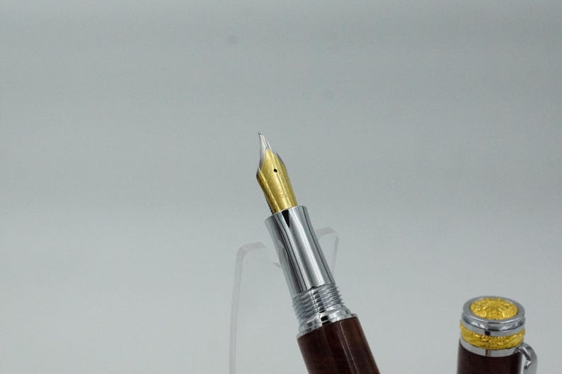 May include: Close-up of a fountain pen with a gold nib and a brown wooden barrel. The pen has a silver metal section and a decorative gold-colored cap. The pen is displayed against a white background.