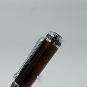 May include: Close-up of a luxury pen with a dark brown, wood-grain barrel and silver accents. The pen features a silver clip and decorative detailing on the cap. The pen's design suggests a high-end writing instrument.