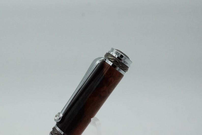 May include: Close-up of a luxury pen with a dark brown, wood-grain barrel and silver accents. The pen features a silver clip and decorative detailing on the cap. The pen's design suggests a high-end writing instrument.