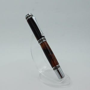 May include: A handcrafted pen with a dark brown and reddish-brown wooden barrel and silver-colored metal accents. The pen is displayed on a clear acrylic stand, showcasing its design. The pen's cap and tip are silver, adding contrast to the wood.