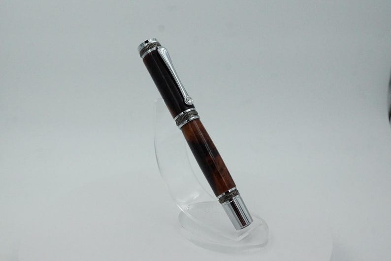 May include: A handcrafted pen with a dark brown and reddish-brown wooden barrel and silver-colored metal accents. The pen is displayed on a clear acrylic stand, showcasing its design. The pen's cap and tip are silver, adding contrast to the wood.