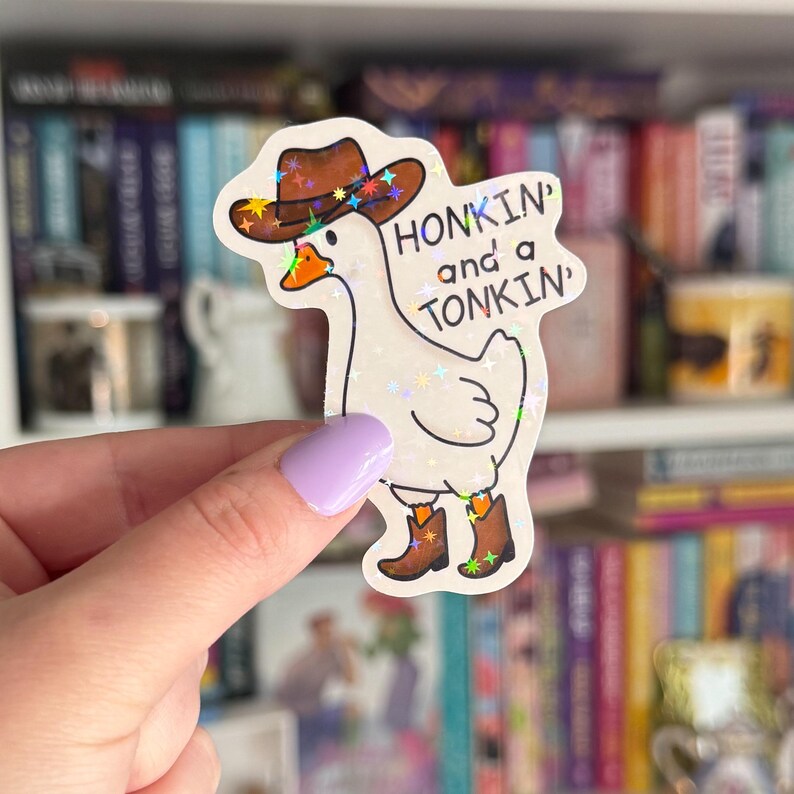 Op de afbeelding: Een witte gans met een bruine cowboyhoed en laarzen met een glinsterende, regenboogkleurige afwerking. De gans houdt een bord vast met de tekst "Honkin' and a Tonkin'".