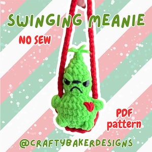 May include: A green crocheted Grinch-like character with a red heart, hanging from red cords. The text "SWINGING MEANIE" and "NO SEW" are displayed above the character. The text "PDF pattern" and "@CRAFTYBAKERDESIGNS" are also visible.